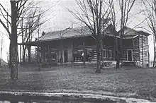 A house of wooden construction with a wrap-around front porch and trees in the front yard