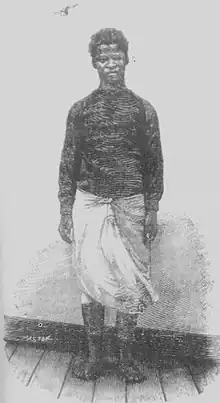 A young boy in a dark top and a white garment tied around his waist standing barefoot in fronf of a blank wall.