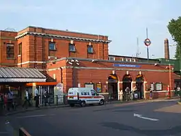 A red-bricked building with a blue sign reading "GOLDERS GREEN STATION" in white letters and people in front all under a blue sky with white clouds