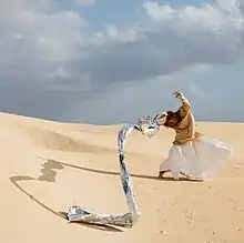 A red headed woman wearing a beige shirt and white skirt stands in the middle of a desert with the wind blowing her arms and a large drapery that flows from her hand.