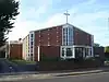 A shallow-roofed building with an extension at the left rear. The façade has small, regularly spaced concrete protrusions, but is mostly brick.  In the centre of this section is a tall 4x5 window extending to the roofline.  A wide, flat-roofed entrance area sits in front of this.  A series of angled ground-to-roof windows run down both sides.