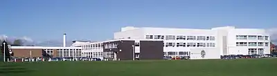 A large group of school buildings. The white building in the background was built in the early 21st century whereas the darker buildings on the left were built in the 1960s or 1970s. In the foreground the front grass field. Since the photo was taken the grass has been replaced by an artificial turf pitch.