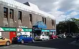 The side entrance of a 1970s brown shopping centre. A post office and charity shop can be seen.