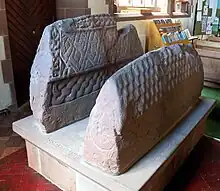 Image 79So-called "Saint's tomb" (left), and "Warrior's tomb" (right), two hogbacks in St. Mary's church, Gosforth. Typically high and narrow Cumbrian style, in the shape of a building with a roof, eaves and walls. The Saint's tomb has a crucifixion on the end, the Warrior's tomb has a procession of armed men (from History of Cumbria)