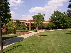 A sidewalk approaches a modern brick and stone building surrounded by trees and greenery.