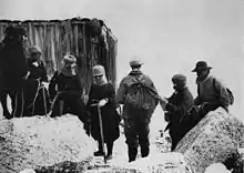 Photo ancienne d'une petite cabane de bois située derrière un groupe de huit personnes chaudement couvertes, avec des rochers au premier plan.