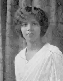 A young, dark-skinned South Asian woman, wearing a white dress. Her dark hair is parted and dressed back and low at the nape. She is looking directly at the camera and not smiling.