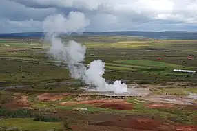 A green landscape with a platform of grey rock. steam is rising from a pool or hole in the middle of the platform