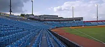 A view from the right field line of the seating bowl at Greer. Blue seats stretch from the right field wall, behind home plate, and beyond the third base dugout.