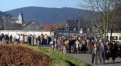 A large number of people of various ages standing and walking along a road in front of a high concrete wall, behind which houses and a church are visible in a wooded valley.