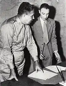 A man in shirt and tie and another wearing a suit stand behind a writing desk. On the wall behind is a map of the Pacific.