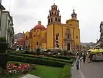 Yellow and red church with cuppolas and two towers one of which has a clock.