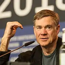 A man wearing a long-sleeved black shirt and watch; his arms and crossed, resting on the back of a chair.