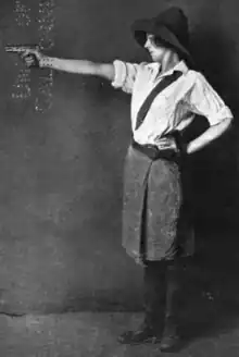 A white woman standing in profile, one arm on hip, other arm outstretched and holding a pistol. She is wearing a hat with a wide brim, a white blouse with sleeves rolled up, and a knee-length skirt with dark tights or boots.