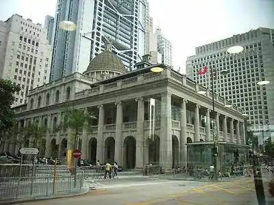 The grey dome and front gable of a granite neo-classical building, with a skyscraper in the background against a clear blue sky