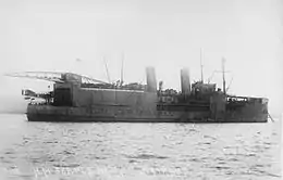 Black and white photograph of a ship. The vessel has two funnels, a large boxy superstructure topped by a crane, and an aircraft on her stern