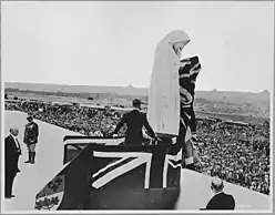 A figure standing on flag covered stage located in front of the statue of Canada Bereft.