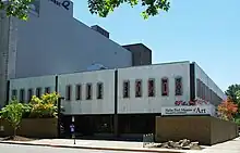 Two-story, marble faced building in with three sections on the second floor, each with tall, rectangular stained glass windows. First floor is slightly recessed and contains metal framed windows covering nearly from ceiling to floor, with entrance near the center. To left and right of entrance is a brick wall that creates a courtyard between the wall and the building.