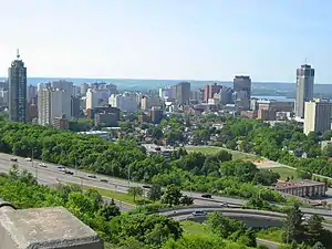 A tree-lined highway is in the foreground, angled diagonally from bottom right to middle left of the image. Buildings are in the centre, and the background is a sky meeting rolling hills in the distance.