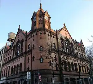 Photograph of a brick building with gables, archways, an octagonal corner tower, and a four-faced clock.