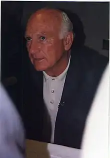 An older, largely bald man with white hair in a blue jacket and white shirt sits while looking up at a person out of frame to the left.