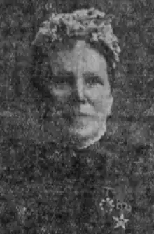 An older white woman with a flower-decorated hat on her head, dressed in a high-collared dark jacket with two badges pinned to the chest