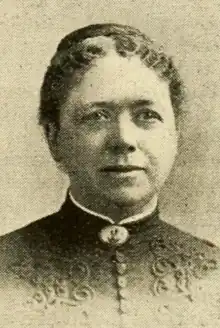 A middle-aged white woman wearing a high-collared buttoned-up dark jacket with braid embellishment and a brooch pinned at the throat; her dark hair is center-parted dressed in a neat updo, and appears to be very wavy