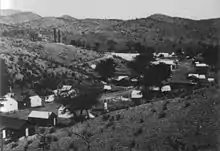 A street of about 30 one-story buildings stretches along the floor of a dry, narrow valley. A few trees grow near the houses, but only low, widely separated desert shrubs grow on the hills above the valley. Two smokestacks belonging to the Hermosa Mill are visible just over the nearest hill.