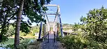 A colored photograph of Hayden Bridge taken from the perspective of someone about to walk onto the bridge. The bridge is shown with its renovations, including a wooden walkway and railing, and is outfitted for use by pedestrians, concealing the underlying railroad beneath.