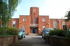 Red brick building with central clock tower. In the foreground are plants and parked cars.