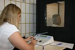 a female medical professional is seated in front of a special sound-proof booth with a glass window, controlling diagnostic test equipment. Inside the booth a middle-aged man can be seen wearing headphones and is looking straight ahead of himself, not at the audiologist, and appears to be concentrating on hearing something