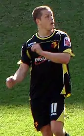 A man in a black football shirt and shorts, with yellow outline, standing on a grass field.