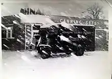 A black and white photo of a truck with a rounded front sitting in the snow in front of a one-story brick building with the words "CANNING SETS" on its outer wall