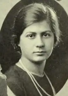 A young white woman, dark hair parted on the side, wearing a dark dress or blouse with a long strand of pearls