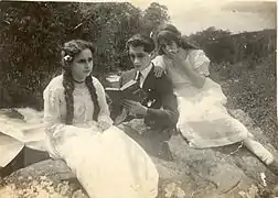 Black and white photo showing a man reading a book, next to two young women sitting on a rock.