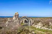 Photograph of a ruined arch supported by metal structures in countryside with open water in the distance.