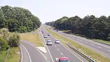 A highway passes beneath the camera and continues straight into the horizon. It is surrounded by forests on either side and contains no guardrail to separate opposite flows of traffic.