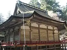 Three-quarter view of a proportionally tall wooden building with a veranda with red hand rail. The corner of the eaves appear cut off.