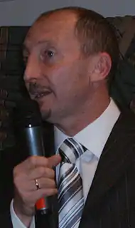 A man wearing a shirt, tie and jacket; holding a microphone.