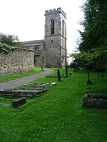 The east end of a stone church is seen through a graveyard from the north. On the right is a battlemented tower and the nave extends to the left