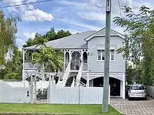 A long shot of a high-set Queenslander residential house made of timber with a corrugated iron roof. An external staircase leads to a veranda on the upper level of the house, and a white picket fence sits at the front of the property.