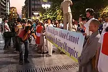 A crowd of protesters in a street displays a banner reading "Human Rights Abuse Cannot Co-exist with Beijing Olympics." Near the center of the image, a photographer holds a camera level with the banner while looking through the viewfinder.