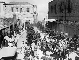 In a black and white photograph, a large group of turbaned men on horseback ride through a dusty, sunlit street and into the distance, obscured by dust. A crown of civilians watch the men pass. A large, brick-built, two-storey building is on the left, and a similar structure is on the right.