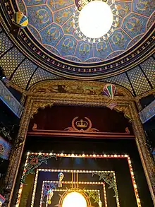 The interior of  a grand theatre with a decorated roof, taken from the perspective of an audience member. You can see floating paper balloons above the seats that are a part of a festive performance.