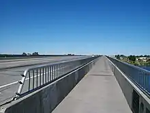 A bridge with metal railings seen from a wide pedestrian path that abuts a freeway