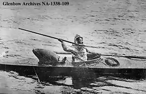 Photo of man sitting in kayak holding spear in throwing position with right arm raised and right hand extended above and behind his head