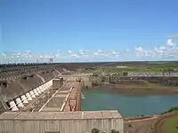 Aerial view of a huge concrete structure located between some low hills and a river, with some large pipes at the base and power lines at the top of the structure and a road