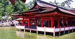 A wooden building with red beams on poles above water.