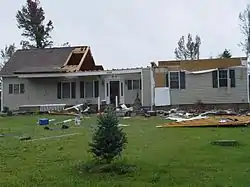 A partially destroyed house, with most of its roof and attic gone, is surrounded by scattered debris.