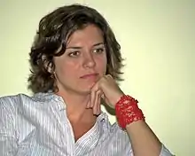 An informal photograph of a young white woman with chin-length blondish hair, wearing a striped button-down shirt and a red bracelet.